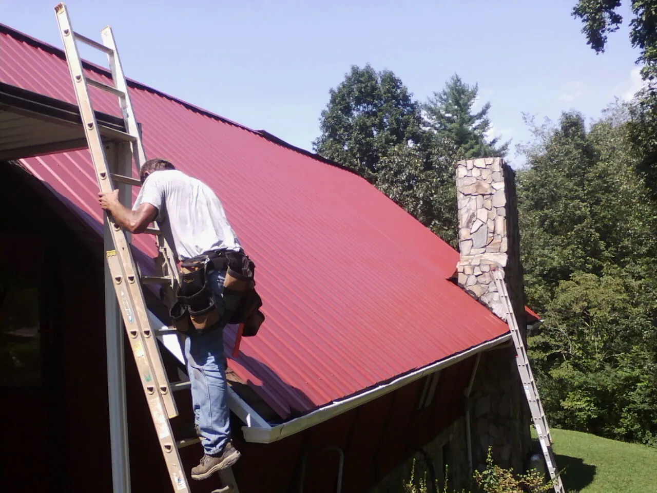 Roofer on a ladder beside a red metal roof and stone chimney — Soffit & Fascia Repair by Roofing Brothers in Dundee